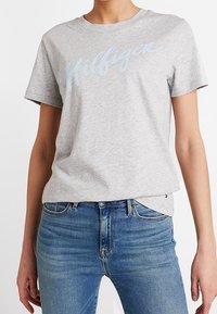 Camiseta de algodón gris con un logo "Hilfiger" en azul claro en el pecho, combinada con jeans de denim azul. Diseño clásico de cuello redondo.