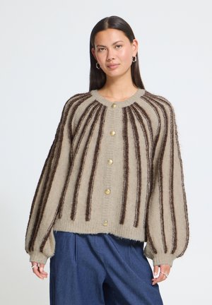 Femme portant un cardigan beige avec des rayures verticales brun foncé et des boutons dorés, associé à un pantalon bleu ample, debout devant un fond blanc.