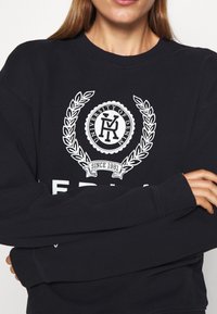Marinblå sweatshirt med vit grafik som visar en cirkulär logotyp och lagerdesign. Ribbad fåll och halsringning, långa ärmar, avslappnad passform.
