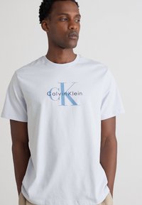 Camiseta de algodón azul claro con un gráfico en la parte frontal que dice "Calvin Klein" en texto azul. Mangas cortas y cuello redondo.