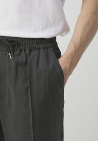 Personne portant un pantalon gris foncé à taille élastique avec cordon, main dans la poche, et une chemise blanche à manches courtes visible à la taille.
