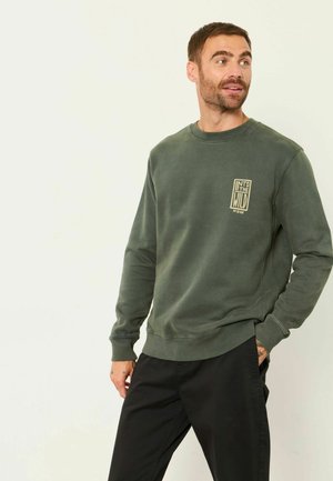 Hombre con sudadera verde de cuello redondo con texto "Into the Wild" y pantalones negros, mirando de lado con una mano en el bolsillo.