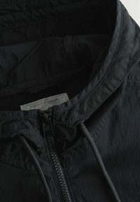 Jachetă ușoară neagră, cu glugă, având un fermoar frontal, șnururi ajustabile și o etichetă care spune "Premium Grade". Textură netedă.