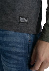 Langærmet grå skjorte med en tekstureret overflade og et mærke med "Petrol Ind" på kanten, parret med blå denim jeans.