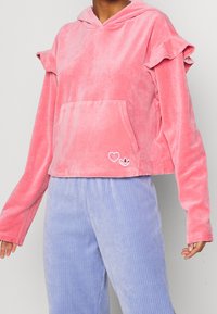 Sudadera de terciopelo rosa con capucha ajustable, mangas con volantes, bolsillo frontal tipo canguro y acentos bordados de corazón y logotipo. Diseño corto.