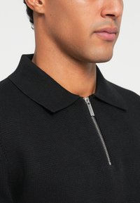 Pullover preto com gola e fecho de zip na frente. Feito de tecido texturizado com detalhes de costura visíveis.