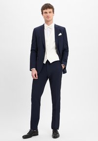 Marineblaues maßgeschneidertes Anzug mit einer doppelt-geknöpften cremefarbenen Weste, weißem Hemd und passender Fliege. Abgerundet mit schwarzen eleganten Schuhen.