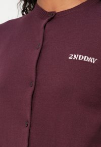 Cardigan lavorato a maglia color bordeaux con collo rotondo, chiusura frontale con bottoni e testo ricamato bianco "2ND DAY" sul lato sinistro. Tessuto morbido.
