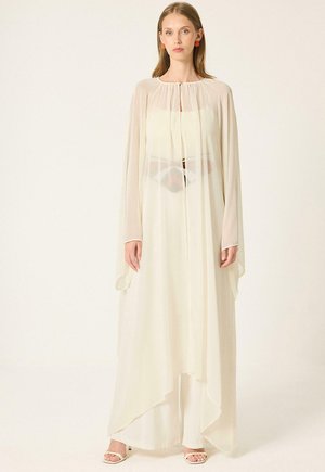 Femme portant une longue robe blanche transparente avec de larges manches et des sandales blanches à talons, debout devant un fond clair uni.