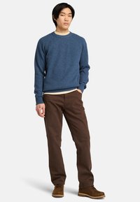 Blå stickad tröja med ribbad textur, rundad halsringning, bärs över en ljus skjorta. Kombinerad med bruna byxor och bruna stövlar.