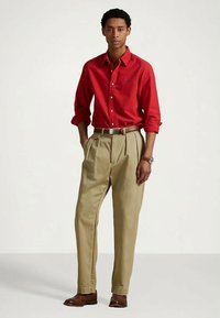 Camisa vermelha de manga comprida com botões, calças plissadas bege, cinto castanho, sapatos castanhos. Fundo de cor clara, modelo está em pé com as mãos nos bolsos.