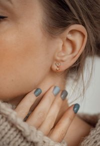 Orecchini a perno dorati con design a cuore e accenti di cristallo trasparente, indossati da una persona con smalto neutro e tessuto lavorato a maglia leggero.
