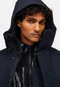 Marinblå isolerad jacka med avtagbar huva, med zippered fram, fleecefoder och zippered fickor med en strukturerad svart mönster.