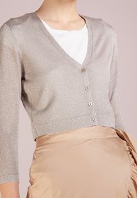 Silverfärgad metallisk kort cardigan med V-ringning, trekvartslånga ärmar och fem knappstängning, bärs över en vit skjorta och en beige kjol.