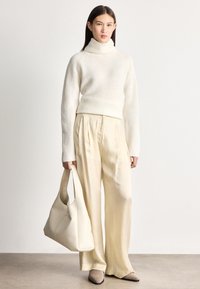 Γυναίκα με κρεμ πουλόβερ ζιβάγκο και σατέν παντελόνια με φαρδιά μπάσκα, κρατώντας μια μεγάλη τσάντα σε ανοιχτό εκρού, φοράει μπεζ παπούτσια με μυτερή μύτη.