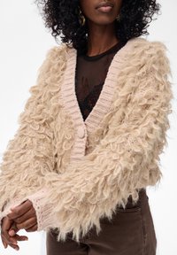 Kobieta w beżowym, teksturowanym kardiganie z dzianiny, nosząca czarną, przezroczystą bluzkę w kwiaty oraz brązowe spodnie, z złotymi kolczykami w kształcie kół.