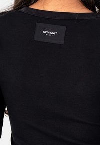 Zwarte geribbelde shirt achterkant met een genaaid zwart etiket met de tekst "SIXTH JUNE PARIS" nabij de halslijn onder lang donker haar.