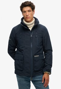 Navy blauwe ritsjack met twee voorzakken, capuchon met koord, en een getextureerde kraag, met merkaccenten op de mouwen en zak.