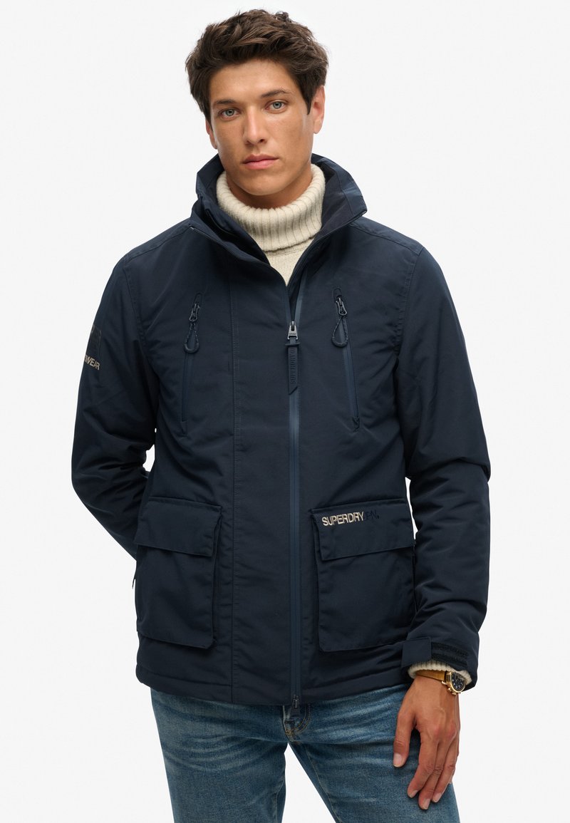 Navy blauwe ritsjack met twee voorzakken, capuchon met koord, en een getextureerde kraag, met merkaccenten op de mouwen en zak.