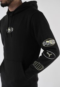 Sudadera negra hecha de material suave, con un logo central y gráficos en las mangas en dorado y blanco. Incluye un bolsillo tipo canguro y capucha con cordón.