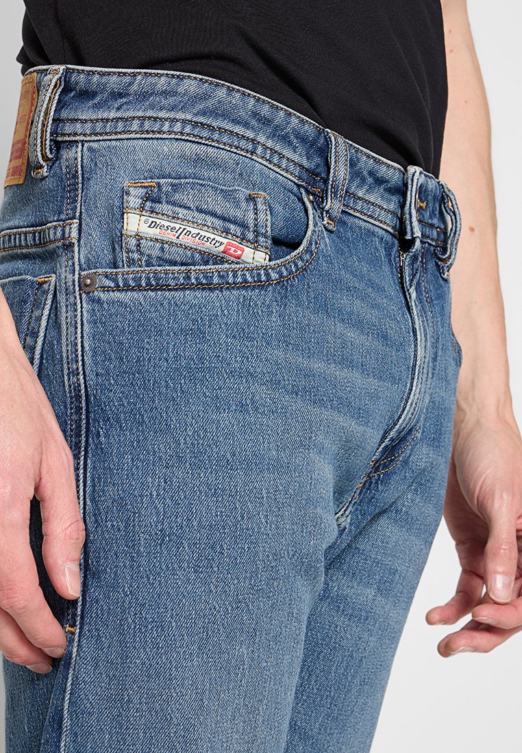 Gros plan sur une personne portant un jean bleu Diesel avec un patch logo visible sur la petite poche avant, et un t-shirt noir rentré.