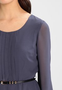 Žena ve tmavě šedých šatech s dlouhým rukávem, které mají vertikální záhyby na hrudi, a s úzkým černým opaskem se zlatou sponou.