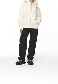 Persona che indossa una felpa crema con zip, pantaloni cargo neri con molte tasche e scarpe da ginnastica nere con lacci bianchi su uno sfondo bianco.