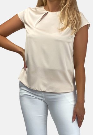 Femme portant un chemisier en satin crème à manches courtes, rentré dans un pantalon blanc ajusté, debout devant un fond uni.
