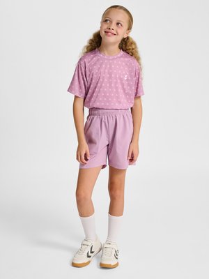 Jong meisje met krullend haar, gekleed in een roze T-shirt met patroon en bijpassende korte broek, witte sokken en witte sneakers, staand en glimlachend.