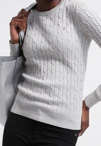 Pull gris clair en maille torsadée avec encolure ronde, poignets et ourlet côtelés. Logo rouge petit sur la poitrine. Bas noir et sac à main gris.