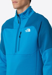 Uomo che indossa una giacca in pile blu brillante con zip, con il logo The North Face sul petto sinistro, colletto alto e tasca a zip sul petto.