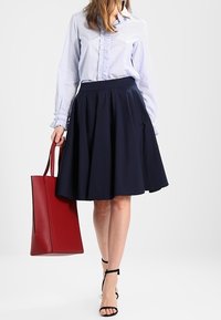 Jupe évasée navy associée à une chemise rayée bleu clair avec des volants, des sandales noires et un sac fourre-tout rouge. Tissu lisse, design décontracté.