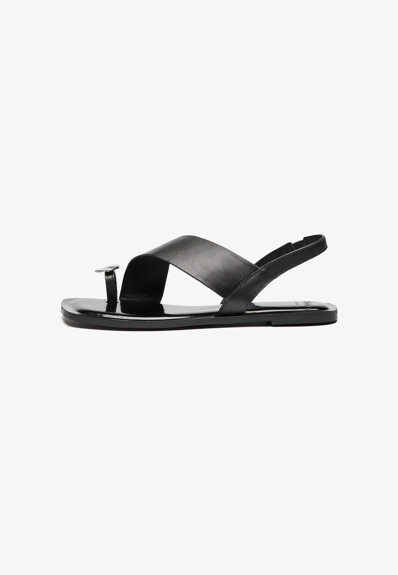 Sandal in pelle nera con un ampio design a incrocio, suola piatta e un elemento rotondo sulla striscia del piede. Texture liscia, stile minimalista.