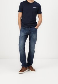 Navy t-shirt with a small graphic on the left chest, paired with blue jeans and brown lace-up sneakers. Simple, casual fit.