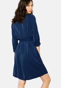 Robe bleu marine avec un col, des manches trois-quarts et une taille ceinturée. Le tissu a une texture lisse et légère.