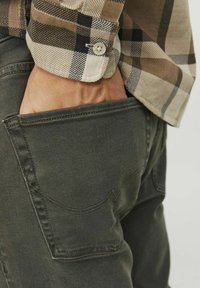 Vaqueros de mezclilla verde con un detalle en el bolsillo trasero y costuras de contraste, combinados con una camisa de cuadros en tonos marrón, negro y beige con puños abotonados.