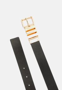 Cinturón de cuero negro con una hebilla de metal en tono dorado con un diseño en capas y nombre de marca grabado. Incluye múltiples agujeros de ajuste.