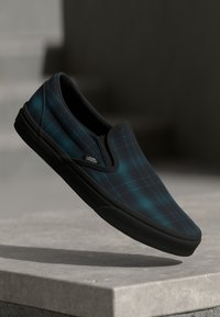 Tenisky Vans slip-on s tmavě modrým a tyrkysovým kostkovaným vzorem, černou gumovou podrážkou a polstrovaným límcem, které mají hladký textilní povrch.
