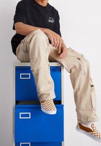 Beige cargo kalhoty se zipy, černé tričko s logem a hnědé kostičkované slip-on boty, sedící na modré zásuvkové jednotce.