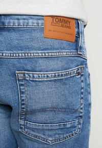 Modré džíny z denimu s koženou nášivkou značky "TOMMY JEANS." Obsahují zadní kapsu s viditelným prošíváním a jemným vyblednutím.