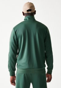 Πράσινο αθλητικό μπουφάν από λείο ύφασμα, με λευκές πλαϊνές ρίγες, ψηλό γιακά, ριμπ μανσέτες και κομψή εφαρμογή.