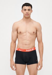 Črne športne hlačke z oranžnim logotipom Nike na pasu. Imajo tesen oprijem, gladko tkanino in kontrastne šive.