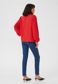 Blouse rouge à manches longues avec une finition texturée et des épaules froncées, associée à un jean skinny bleu et des mules noires à talons. Fond uni.