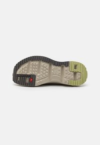 La semelle de la chaussure présente un design multi-texturé avec une combinaison de couleurs noir et beige clair, un accent vert au talon et des crampons de traction proéminents.