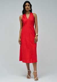 Vestido de lino rojo a media longitud con un escote en V profundo, diseño sin mangas y silueta en A. Lleva unos tacones plateados de tiras y acentos de joyería.