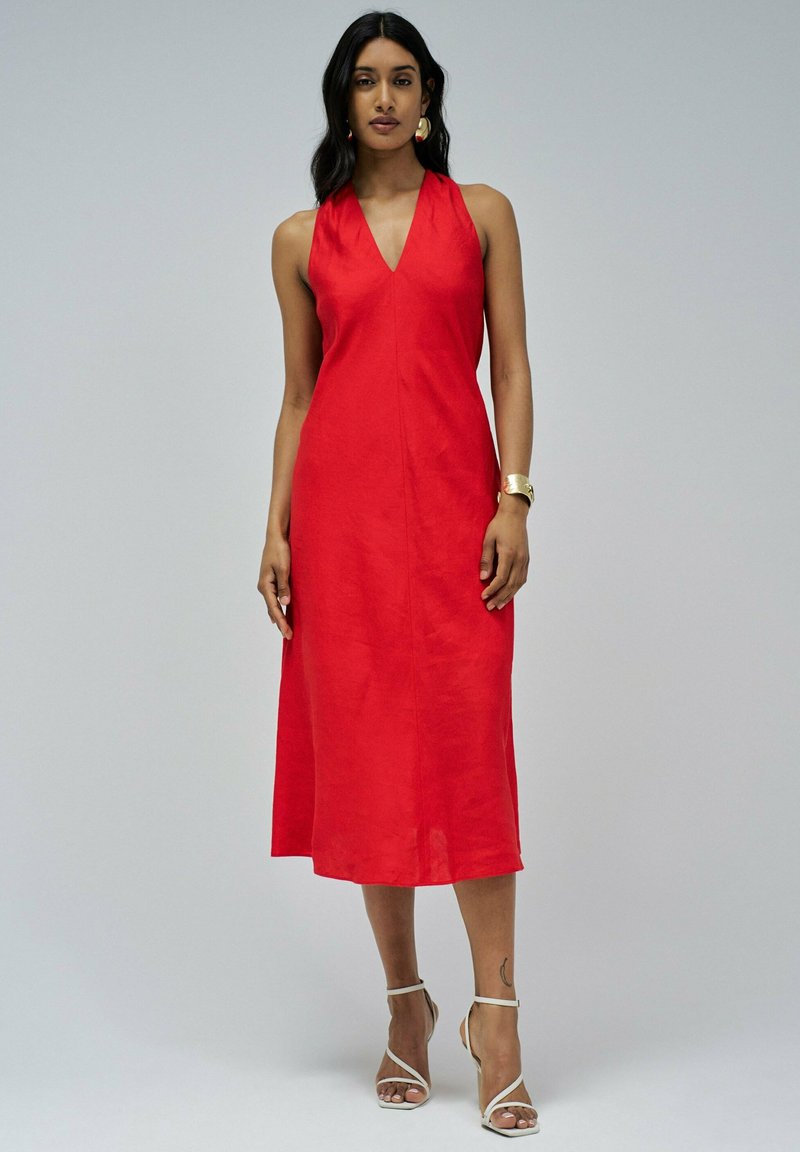 Vestido de lino rojo a media longitud con un escote en V profundo, diseño sin mangas y silueta en A. Lleva unos tacones plateados de tiras y acentos de joyería.