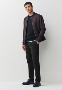 Veste bomber marron foncé portée sur un pull col rond noir, associée à un pantalon noir et des chaussures noires. Couleurs unies, style décontracté, détails minimalistes.