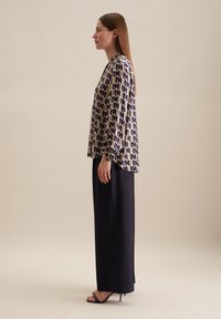 Blouse ample avec un imprimé géométrique en marine et crème, associée à un pantalon long sombre. La blouse a des manches longues et un col haut.