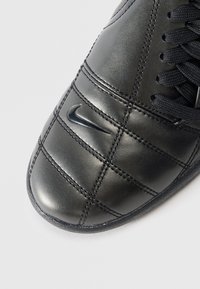 Black leather Nike sneaker with quilted stitching and black laces, showing the toe and part of the side on a white background.