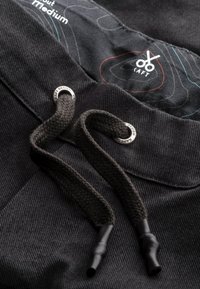 Pantalons noirs avec cordons tressés et œillets en métal. Le tissu présente un motif texturé. L'intérieur comporte un design graphique en rouge et bleu.